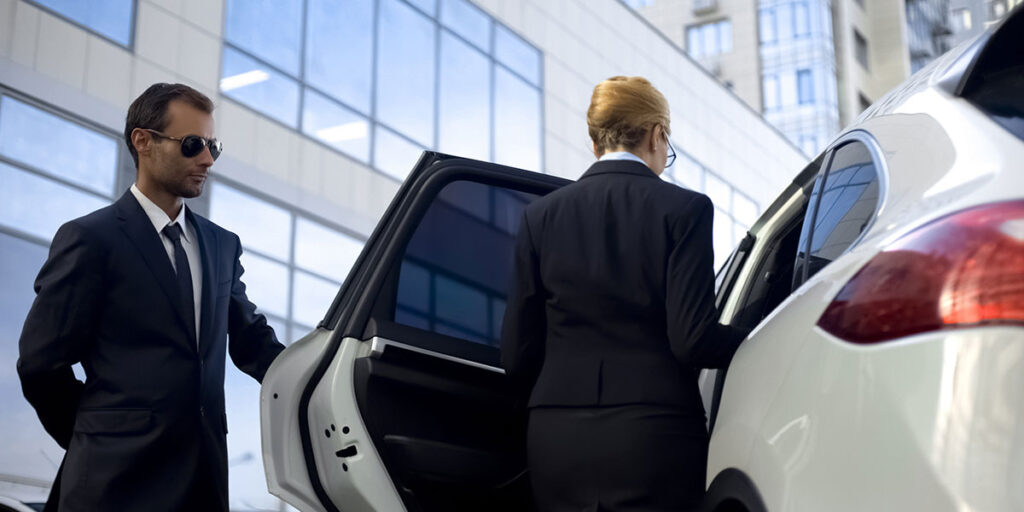 A professional security agent in a dark suit and sunglasses stands beside an open car door as a woman in business attire enters a white vehicle.