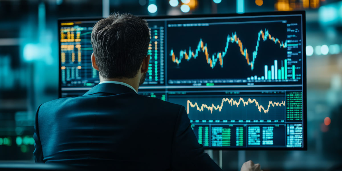 A man in a suit sits at a workstation with multiple large monitors displaying complex data visualizations and line graphs.