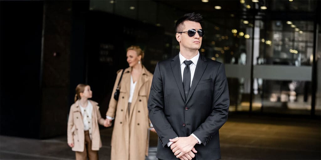 Professional bodyguard in a black suit and sunglasses stands in front of a building, while a woman and young girl in coats walk behind him, holding hands.