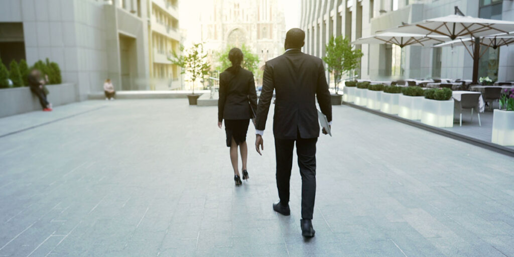 A professionally dressed man and woman walk away down a modern, upscale urban plaza surrounded by tall office buildings.