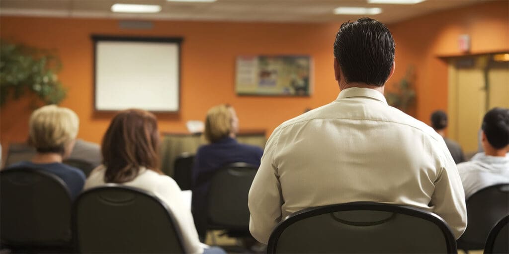 Employees seated in a training room attentively listening to a workplace safety presentation, with a projector screen at the front and an orange-colored wall in the background.