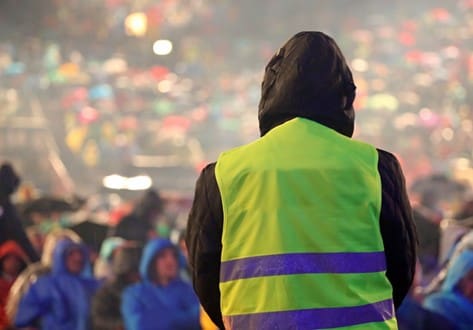 A security personnel wearing a high-visibility vest monitors a large crowd at an outdoor event.