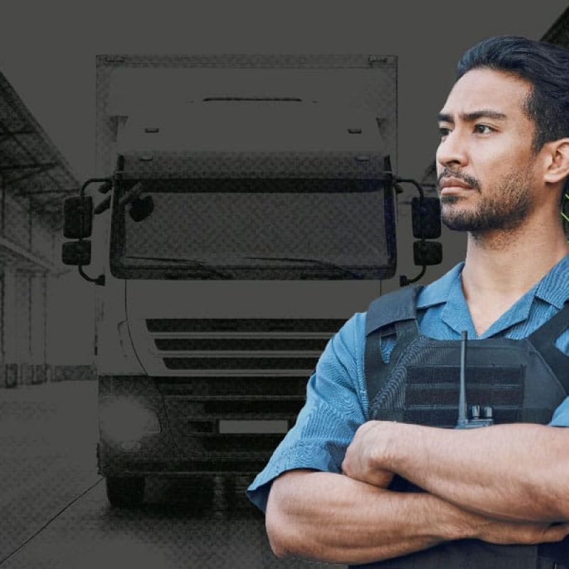 2025-0310_BLOG_HighValueTransport_600x1200 Security officer in uniform and protective vest standing with arms crossed in front of a large truck at a warehouse facility