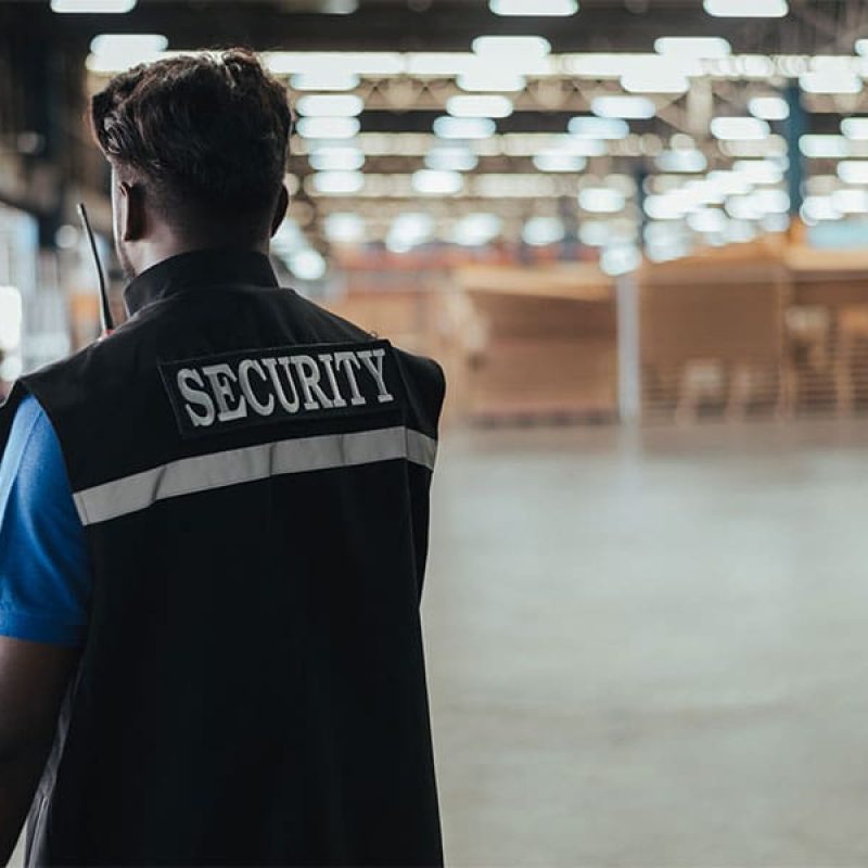 2025-0637_BLOG_HolidaySurge_600x1200 Security guard wearing a vest labeled ‘SECURITY’ patrols a large warehouse