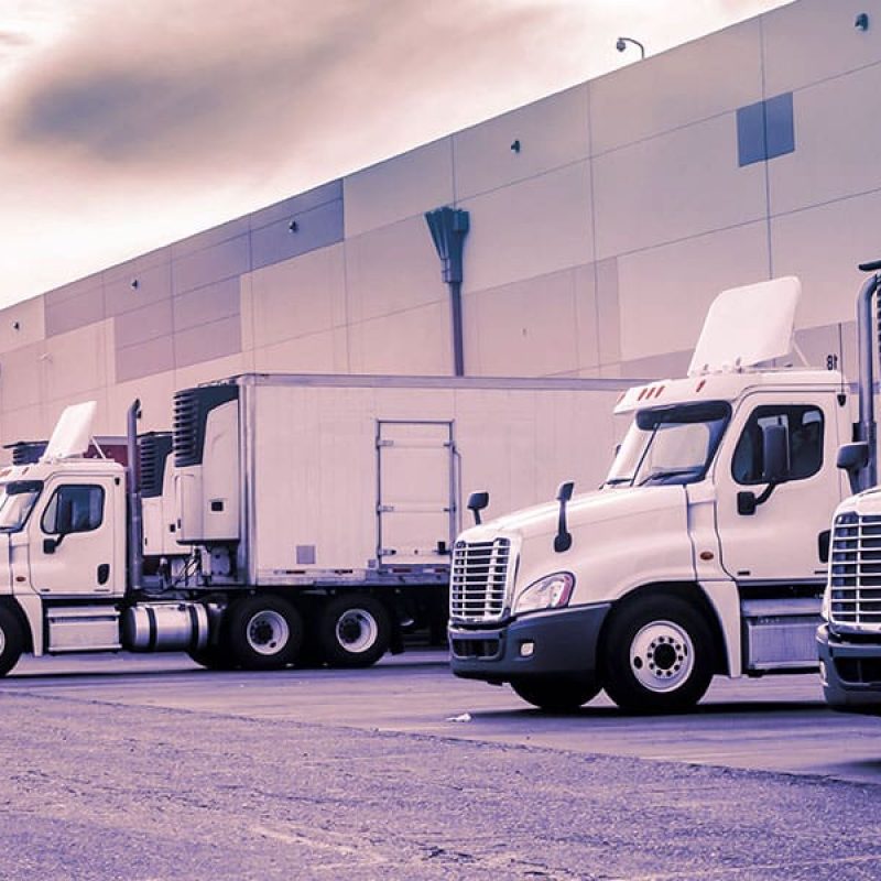 2025-0640_BLOG_CornerstoneHolidaySecurity_600x1200 Row of white semi-trucks parked outside a large warehouse distribution center